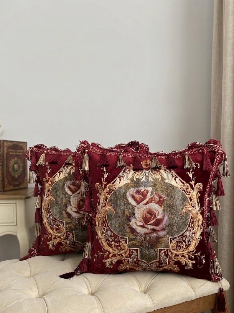 May include: Two decorative throw pillows with a burgundy background and a floral rose design. The pillows have gold trim and tassels. The pillows are placed on a cream-colored bench.
