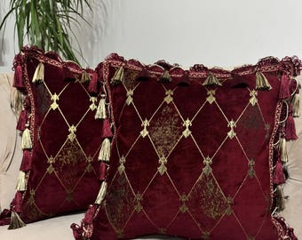 May include: Two burgundy velvet throw pillows with a gold diamond pattern and gold tassels. The pillows have a decorative trim and are placed on a beige surface. The pillows add a touch of luxury to any living space.