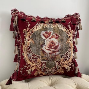 May include: A decorative burgundy pillow with a floral rose design. The pillow features a detailed tapestry with gold accents and is trimmed with tassels. The pillow is placed on a cream-colored upholstered surface.
