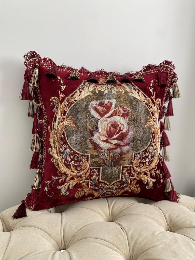 May include: A decorative burgundy pillow with a floral rose design. The pillow features a detailed tapestry with gold accents and is trimmed with tassels. The pillow is placed on a cream-colored upholstered surface.
