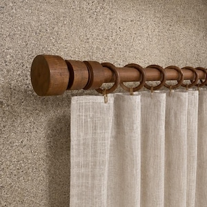 May include: A wooden curtain rod with a cylindrical finial and matching circular rings, supporting a light beige, semi-sheer fabric curtain. The rod and rings feature a smooth, medium-brown wood grain finish, while the curtain displays a visible, woven texture.