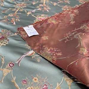 May include: Two pieces of fabric with a floral and bird pattern. One fabric is a muted teal color, and the other is a reddish-brown. The design includes flowers, birds, and ornate branches. A tag is visible on the teal fabric.