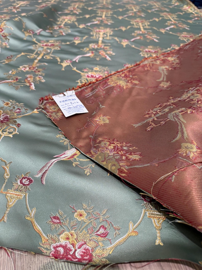 May include: Two pieces of fabric with a floral and bird pattern. One fabric is a muted teal color, and the other is a reddish-brown. The design includes flowers, birds, and ornate branches. A tag is visible on the teal fabric.