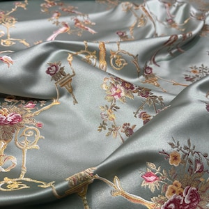 May include: Silky, sage green fabric with a floral and bird pattern. The design features bouquets of pink, yellow, and burgundy flowers, along with gold-colored ornate accents. The fabric has a subtle sheen, creating a luxurious appearance.