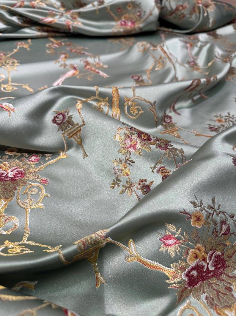 May include: Silky, sage green fabric with a floral and bird pattern. The design features bouquets of pink, yellow, and burgundy flowers, along with gold-colored ornate accents. The fabric has a subtle sheen, creating a luxurious appearance.