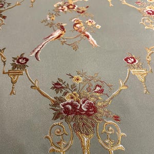 May include: Close-up of a fabric with an ornate design. The fabric has a pale green background with a repeating pattern of gold-colored floral arrangements, birds, and decorative elements. The flowers are primarily burgundy and yellow.
