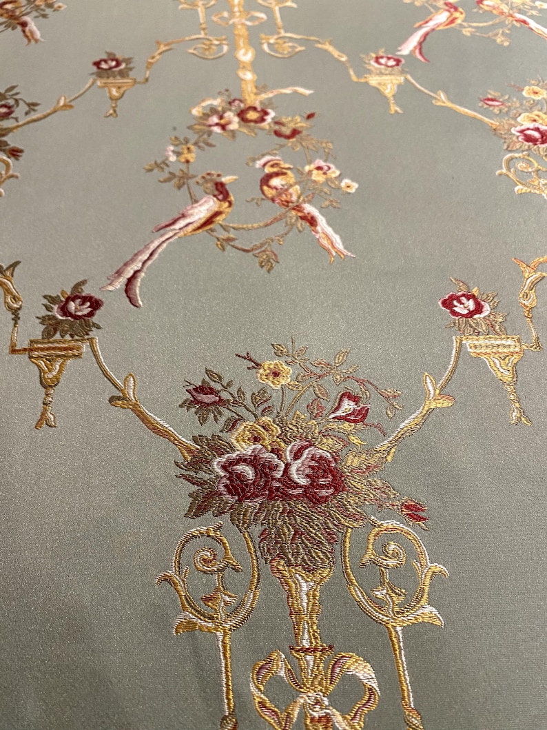 May include: Close-up of a fabric with an ornate design. The fabric has a pale green background with a repeating pattern of gold-colored floral arrangements, birds, and decorative elements. The flowers are primarily burgundy and yellow.