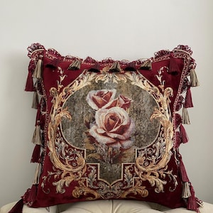 May include: A decorative square pillow in deep red velvet features a central floral design of two roses. The pillow is framed with ornate gold detailing and adorned with matching tassels along the edges, creating a luxurious aesthetic.