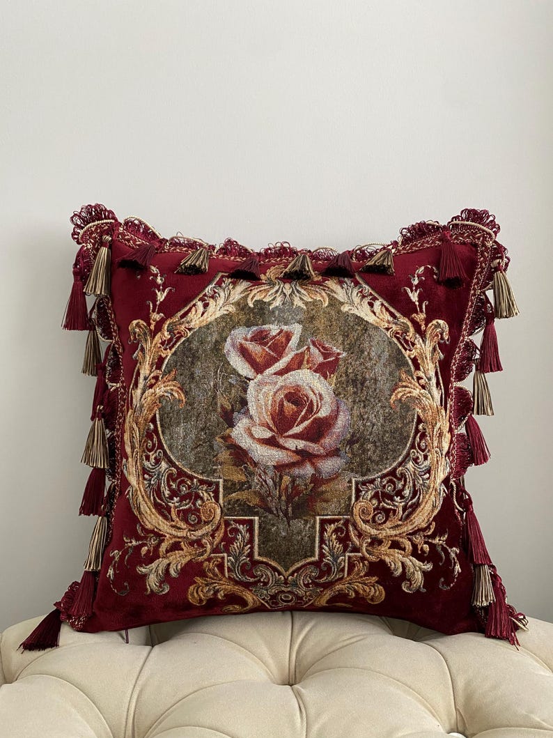 May include: A decorative square pillow in deep red velvet features a central floral design of two roses. The pillow is framed with ornate gold detailing and adorned with matching tassels along the edges, creating a luxurious aesthetic.