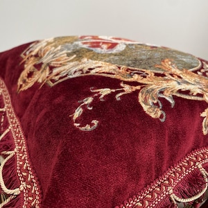 May include: Close-up of a burgundy velvet pillow with ornate gold and beige floral embroidery. The pillow has a decorative fringe trim along the edges, adding a touch of elegance. The design is reminiscent of classic, vintage decor.
