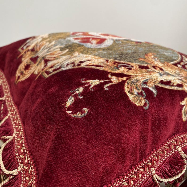 May include: Close-up of a burgundy velvet pillow with ornate gold and beige floral embroidery. The pillow has a decorative fringe trim along the edges, adding a touch of elegance. The design is reminiscent of classic, vintage decor.