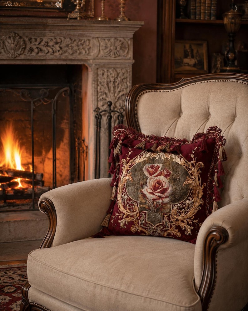 May include: A decorative burgundy pillow with a floral rose design and gold accents rests on a beige armchair. The pillow features tassels and is placed in front of a fireplace with a burning fire.