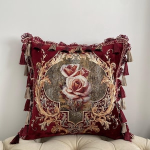 May include: A decorative burgundy throw pillow with a floral rose design. The pillow features a detailed embroidered image of two roses in a central oval frame, surrounded by ornate gold filigree and tassels.