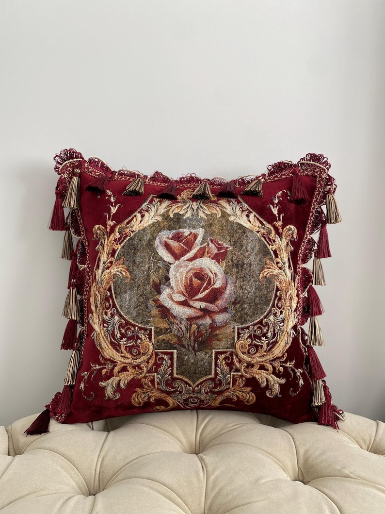 May include: A decorative burgundy throw pillow with a floral rose design. The pillow features a detailed embroidered image of two roses in a central oval frame, surrounded by ornate gold filigree and tassels.