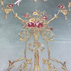 May include: Close-up of a teal fabric with an ornate embroidered design. The design features gold, red, and white floral arrangements, birds, and decorative elements. The fabric has a subtle sheen.