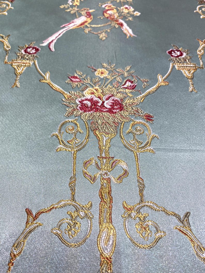 May include: Close-up of a teal fabric with an ornate embroidered design. The design features gold, red, and white floral arrangements, birds, and decorative elements. The fabric has a subtle sheen.