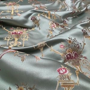 May include: Close-up of a luxurious, sage green fabric with an ornate floral pattern. The design features detailed gold accents and bouquets of red, pink, and yellow flowers. The fabric has a subtle sheen, suggesting a satin or silk material.