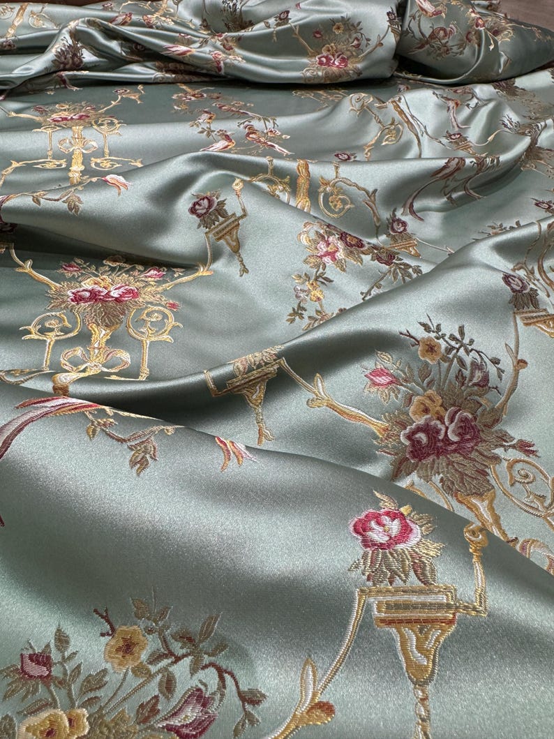 May include: Close-up of a luxurious, sage green fabric with an ornate floral pattern. The design features detailed gold accents and bouquets of red, pink, and yellow flowers. The fabric has a subtle sheen, suggesting a satin or silk material.