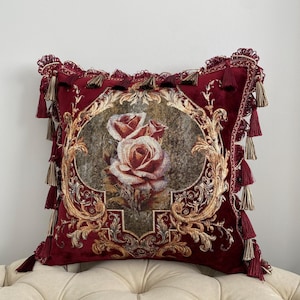May include: A decorative burgundy throw pillow with a floral rose design. The pillow features a detailed gold and brown ornate frame around the roses. Tassels and fringe adorn the edges of the pillow, adding a touch of elegance.
