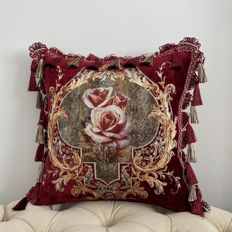 May include: A decorative burgundy throw pillow with a floral rose design. The pillow features a detailed gold and brown ornate frame around the roses. Tassels and fringe adorn the edges of the pillow, adding a touch of elegance.