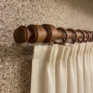 May include: A wooden curtain pole with decorative rings and finials. The pole is a warm brown colour, and the curtains are a light beige. The pole is mounted on a textured wall.