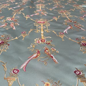 May include: A close-up of a fabric with an embroidered design. The fabric is a muted teal color, with a repeating pattern of gold, red, and white birds, flowers, and ornate branches. The embroidery is detailed and adds texture to the fabric.