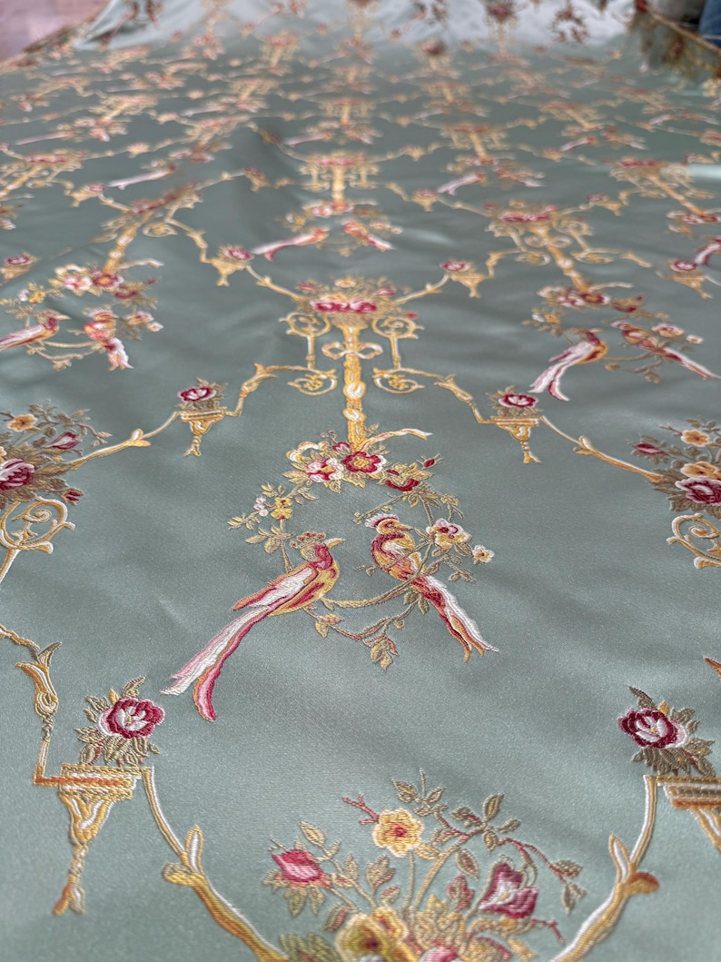 May include: A close-up of a fabric with an embroidered design. The fabric is a muted teal color, with a repeating pattern of gold, red, and white birds, flowers, and ornate branches. The embroidery is detailed and adds texture to the fabric.