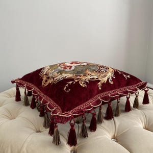 May include: A square, burgundy velvet pillow with an ornate floral design in the center. The pillow is trimmed with gold-colored scalloped edging and burgundy tassels. The pillow is resting on a cream-colored, button-tufted ottoman.