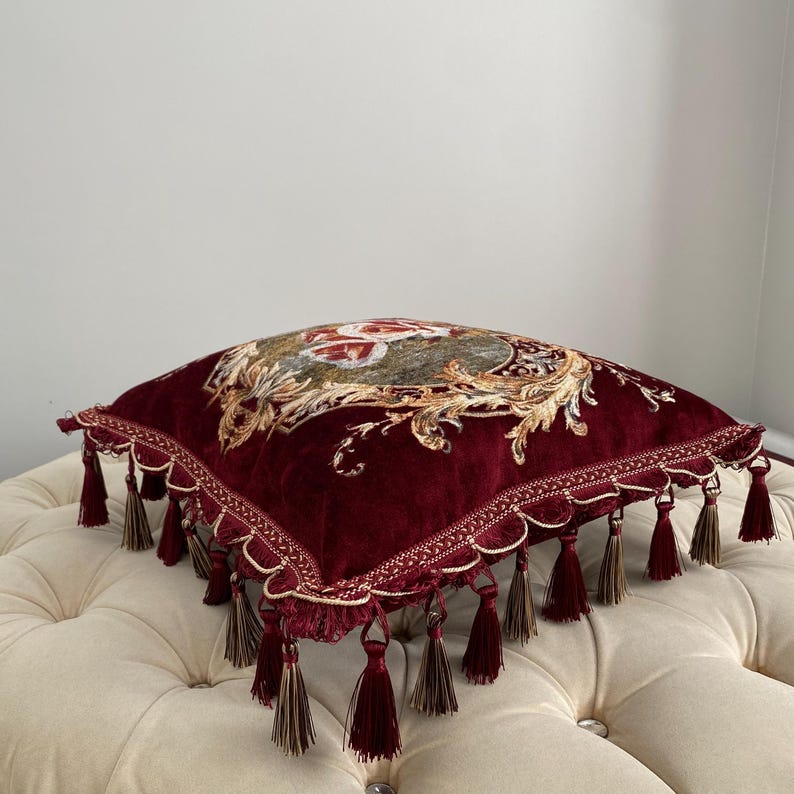 May include: A square, burgundy velvet pillow with an ornate floral design in the center. The pillow is trimmed with gold-colored scalloped edging and burgundy tassels. The pillow is resting on a cream-colored, button-tufted ottoman.