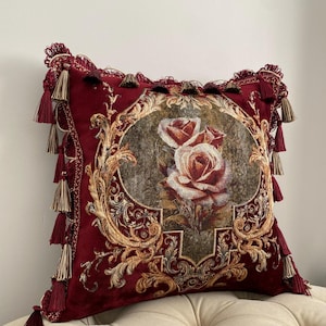 May include: A decorative burgundy pillow with a floral rose design. The pillow features gold-colored ornate detailing and tassels. The center of the pillow displays a detailed image of two roses. The pillow is approximately 18 inches square.