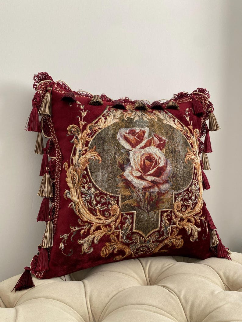 May include: A decorative burgundy pillow with a floral rose design. The pillow features gold-colored ornate detailing and tassels. The center of the pillow displays a detailed image of two roses. The pillow is approximately 18 inches square.