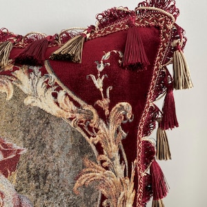 May include: A decorative burgundy pillow with ornate gold and cream-colored floral tapestry. The pillow features a velvet texture, accented with burgundy and gold tassels and trim, creating a luxurious aesthetic.
