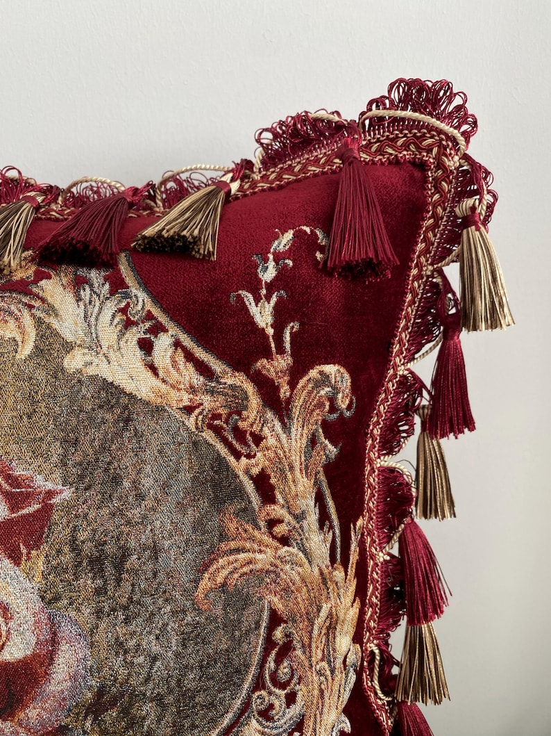 May include: A decorative burgundy pillow with ornate gold and cream-colored floral tapestry. The pillow features a velvet texture, accented with burgundy and gold tassels and trim, creating a luxurious aesthetic.