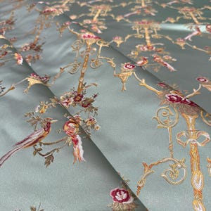 May include: A folded section of fabric with an intricate floral and bird design. The fabric has a pale green background with embroidered details in gold, red, and brown. The pattern includes stylized trees, birds, and floral elements.