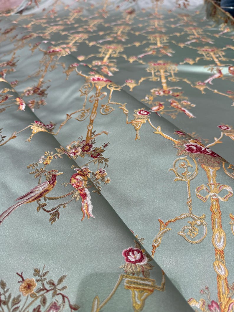 May include: A folded section of fabric with an intricate floral and bird design. The fabric has a pale green background with embroidered details in gold, red, and brown. The pattern includes stylized trees, birds, and floral elements.
