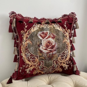 May include: A decorative burgundy throw pillow with a floral design. The pillow features a central oval image of roses in shades of red and cream, surrounded by ornate gold filigree. The edges are trimmed with tassels.