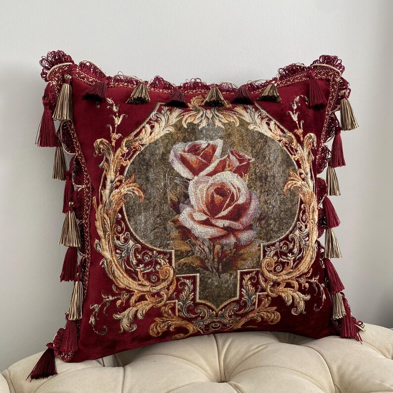 May include: A decorative burgundy throw pillow with a floral design. The pillow features a central oval image of roses in shades of red and cream, surrounded by ornate gold filigree. The edges are trimmed with tassels.