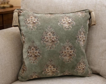 May include: A square decorative pillow with a sage green background and a repeating floral pattern in shades of beige and brown. The pillow has a matching tassel on each corner. The pillow is placed on a beige sofa.