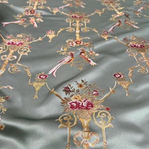 May include: A close-up of a sage green fabric with an embroidered floral and bird pattern. The design features gold-colored candelabras, bouquets of red and yellow flowers, and pairs of birds with long tail feathers.