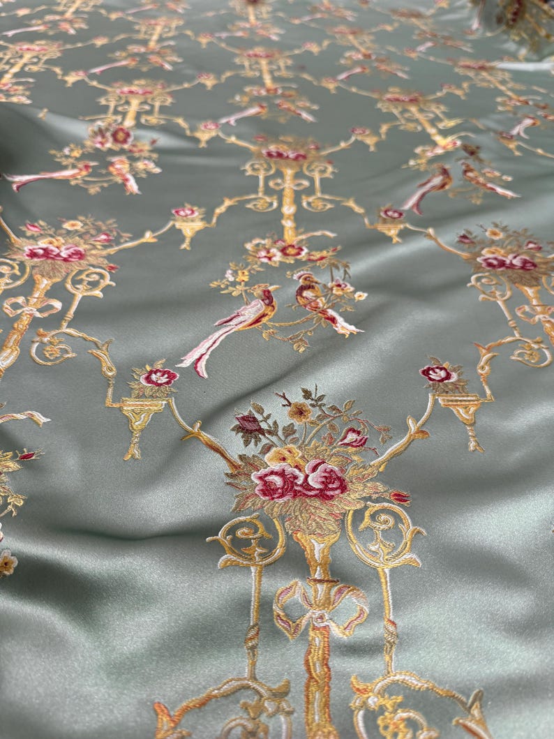 May include: A close-up of a sage green fabric with an embroidered floral and bird pattern. The design features gold-colored candelabras, bouquets of red and yellow flowers, and pairs of birds with long tail feathers.