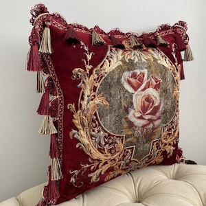 May include: A decorative burgundy pillow with a floral rose design in the center. The pillow features gold filigree and is trimmed with tassels. The pillow is a square shape.