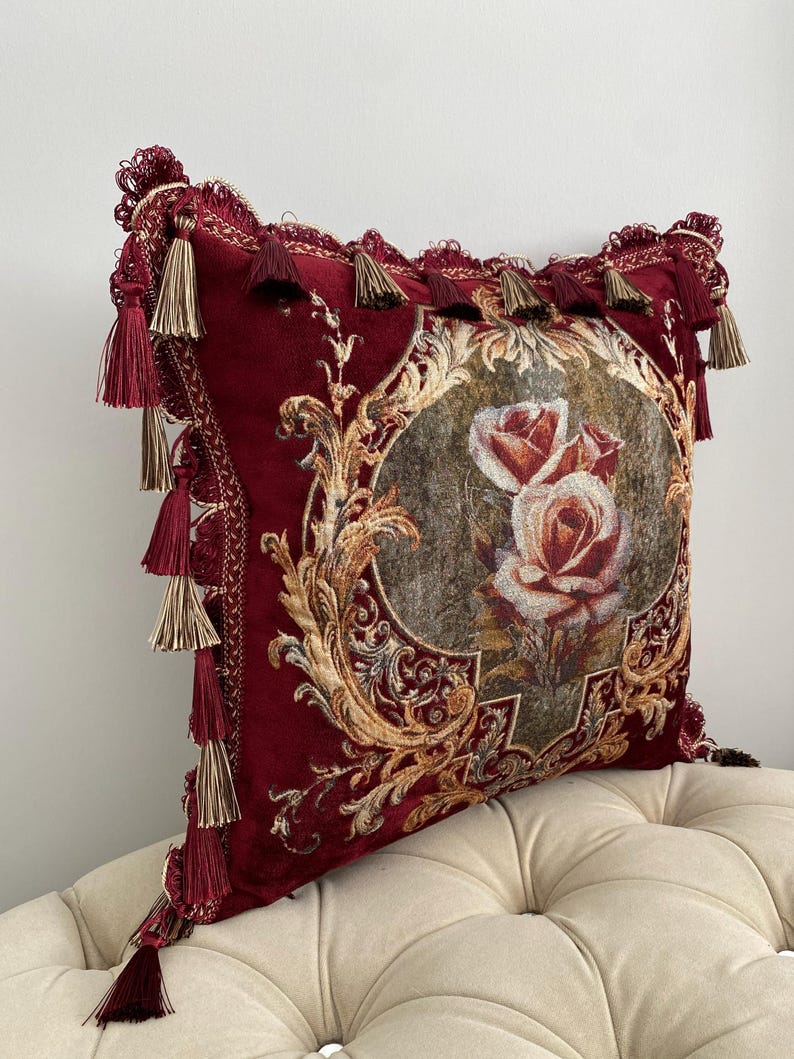 May include: A decorative burgundy pillow with a floral rose design in the center. The pillow features gold filigree and is trimmed with tassels. The pillow is a square shape.