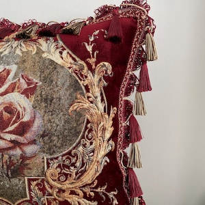 May include: Close-up of a decorative pillow with a burgundy velvet texture. The pillow features a floral design with gold accents and ornate detailing. Burgundy and gold tassels adorn the edges, adding a touch of elegance to the home decor item.