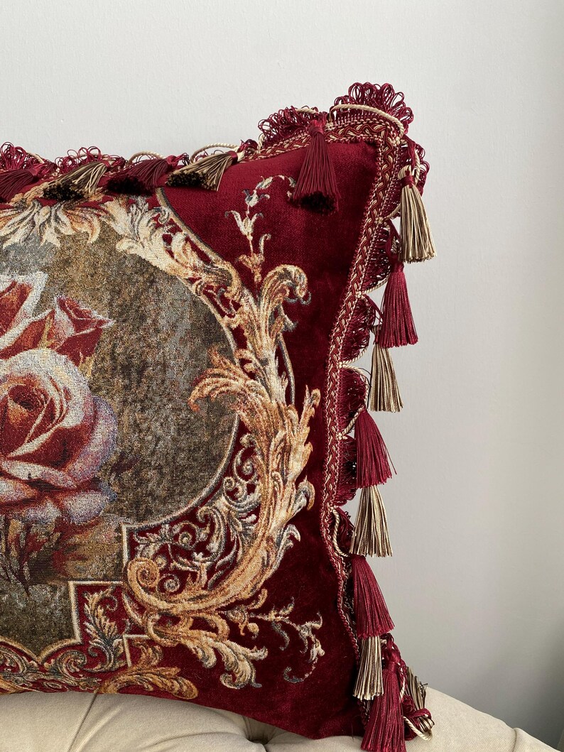 May include: Close-up of a decorative pillow with a burgundy velvet texture. The pillow features a floral design with gold accents and ornate detailing. Burgundy and gold tassels adorn the edges, adding a touch of elegance to the home decor item.