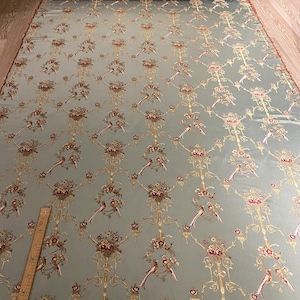 May include: A large roll of fabric with a repeating pattern of birds, flowers, and ornate gold-colored scrollwork on a pale blue-green background. The fabric is rolled up at the top, revealing a brown floral pattern. A wooden ruler is visible.