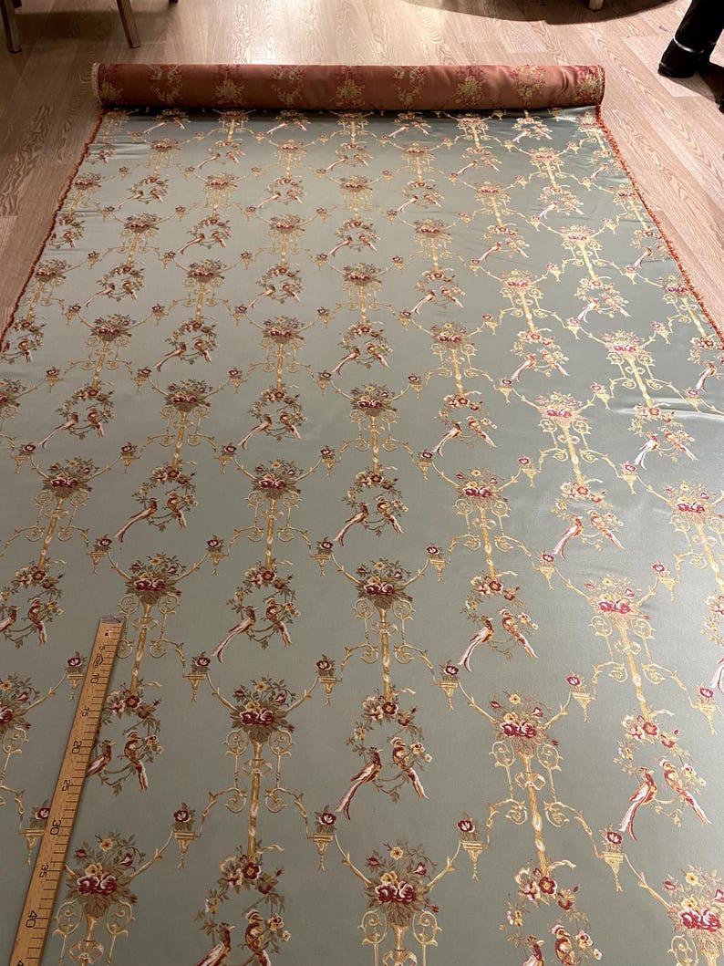 May include: A large roll of fabric with a repeating pattern of birds, flowers, and ornate gold-colored scrollwork on a pale blue-green background. The fabric is rolled up at the top, revealing a brown floral pattern. A wooden ruler is visible.