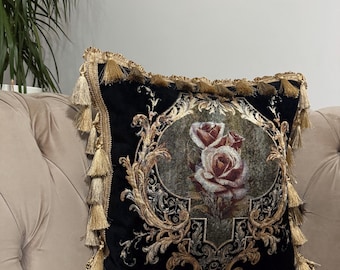 May include: Black decorative throw pillow with a floral rose design in the center. The pillow has gold tassels on the edges and a gold ornate border. The pillow is on a beige sofa.