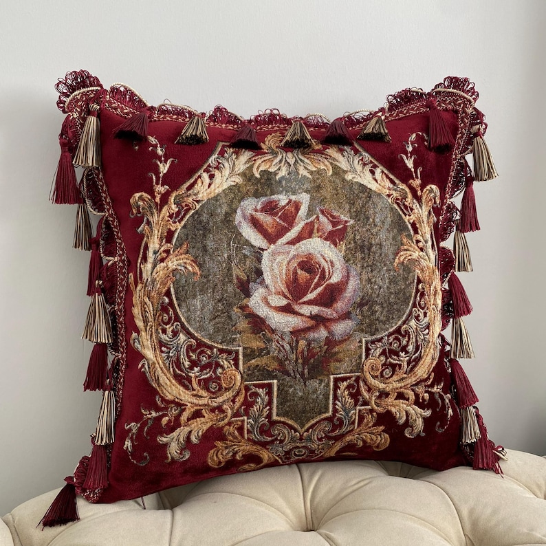 May include: A decorative burgundy throw pillow with a floral design. The pillow features a central oval image of roses in shades of red and cream, surrounded by ornate gold detailing. Burgundy tassels and trim complete the design.