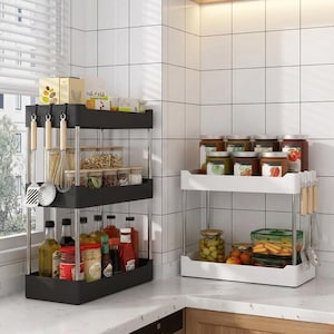 May include: Two-tier kitchen storage shelves, one black and one white, with stainless steel supports. The shelves hold jars, bottles, and utensils. The black shelf has hanging utensils. The white shelf has jars of food.