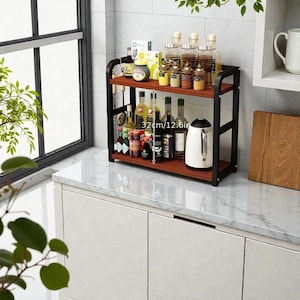 Puede incluir: Estantería de cocina de dos niveles con marco de metal negro y estantes de madera marrón. La estantería mide 32 cm de ancho y contiene varias botellas, tarros y una tetera eléctrica blanca. Sobre una encimera blanca.